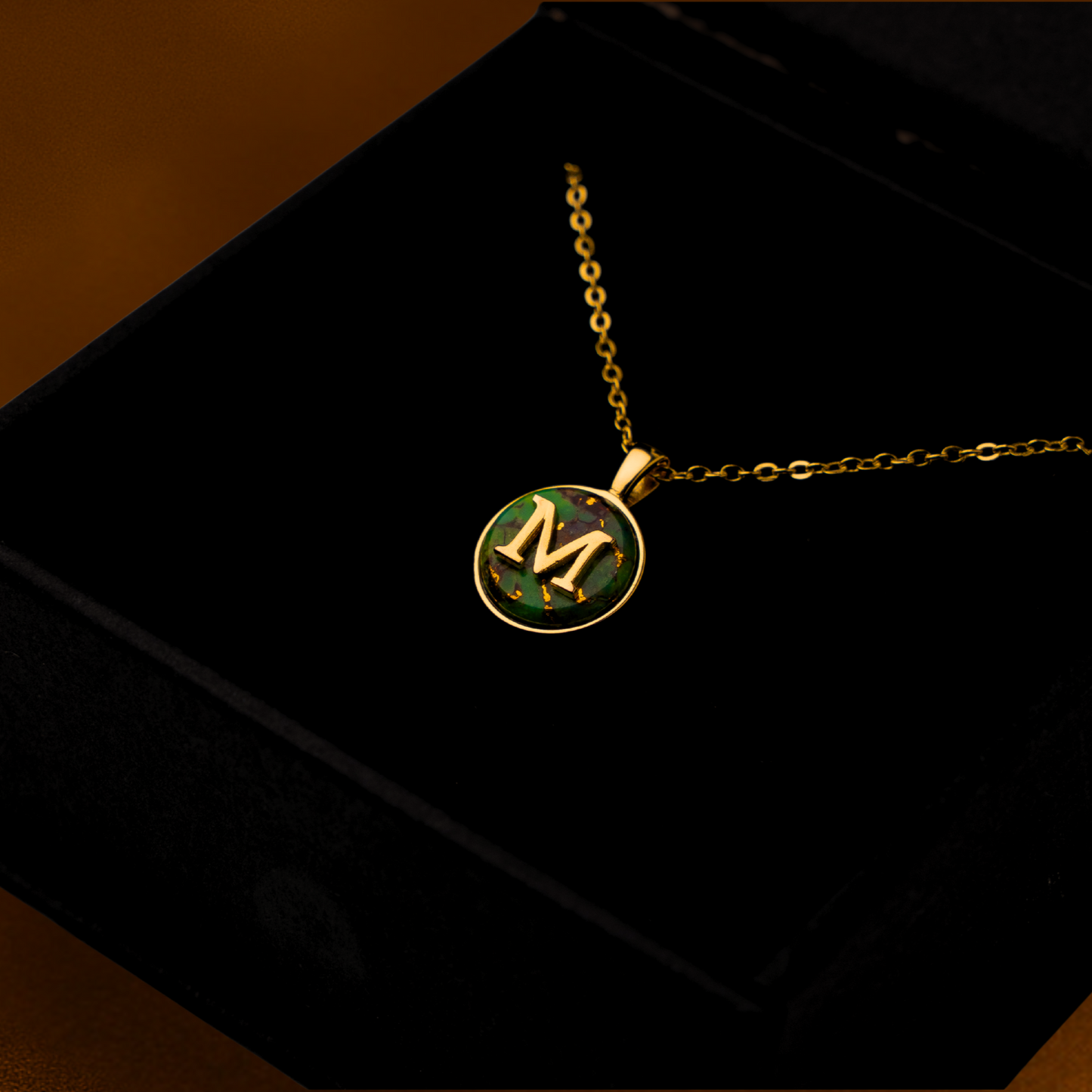 Gold necklace with a circular pendant featuring a letter 'M' on a black box.